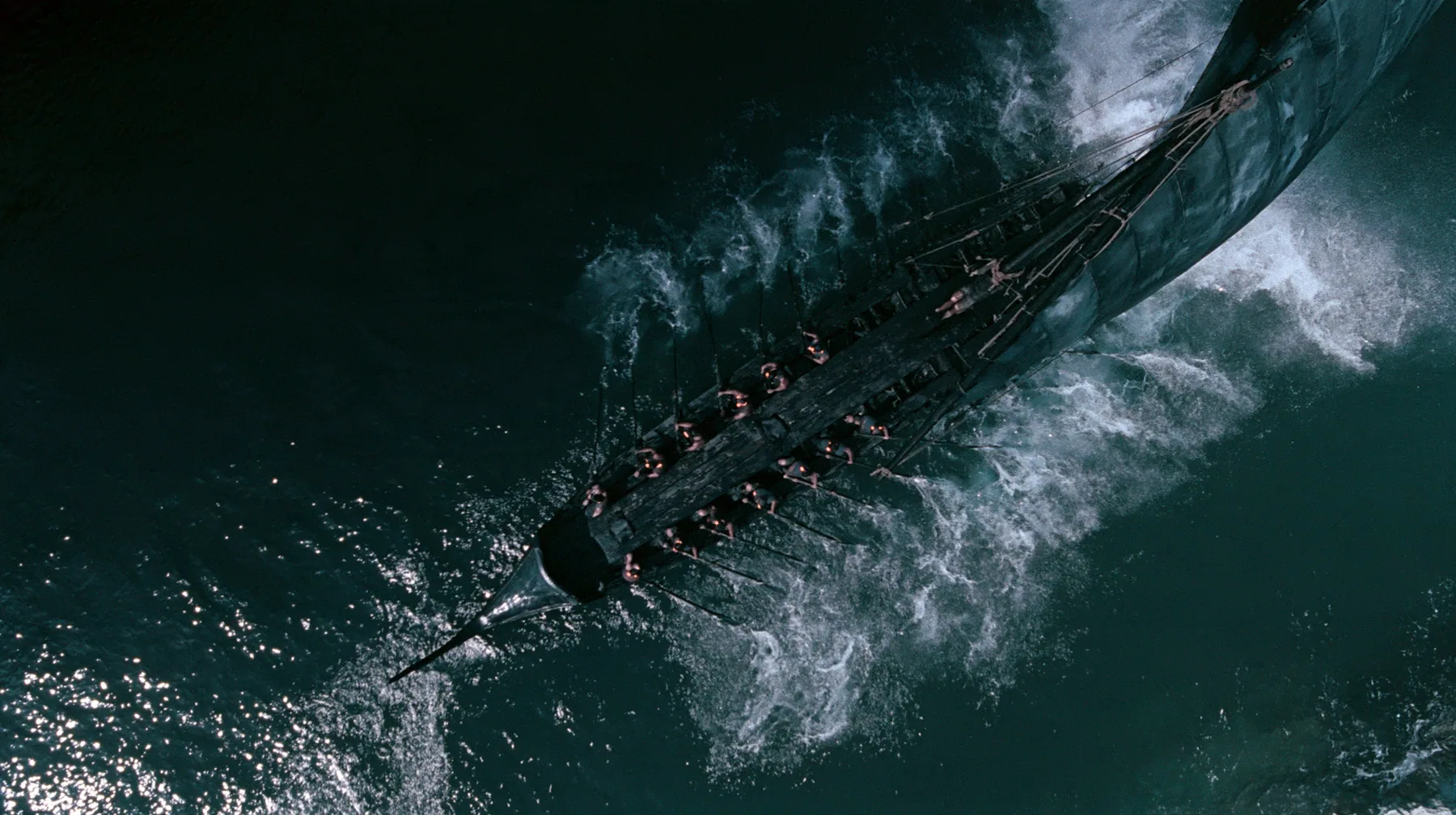 straight-down aerial of a long black ancient Greek galley with a single mast, a square sail and two banks of oars in a narrow strait, half the benches empty, the remaining rowers rowing with orange earplugs, one man…