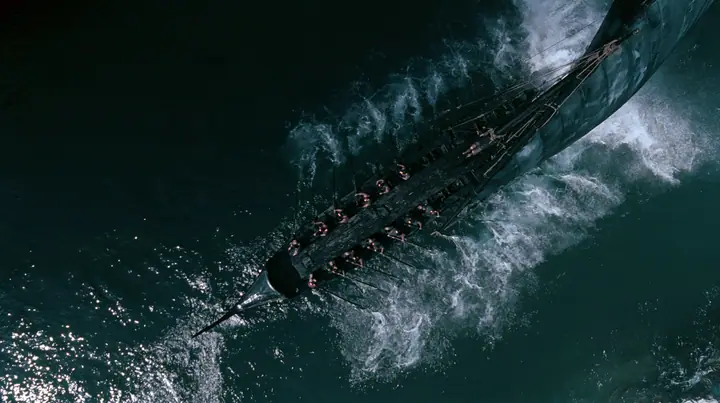 straight-down aerial of a long black ancient Greek galley with a single mast, a square sail and two banks of oars in a narrow strait, half the benches empty, the remaining rowers rowing with orange earplugs, one man…