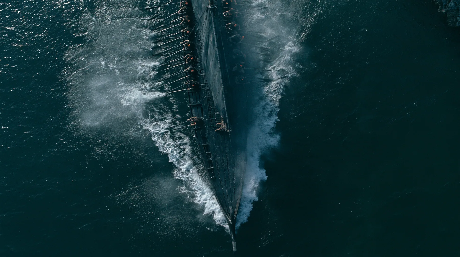 straight-down aerial of a long black ancient Greek galley with a single mast, a square sail and two banks of oars in a narrow strait, half the benches empty, the remaining rowers rowing with orange earplugs, one man…