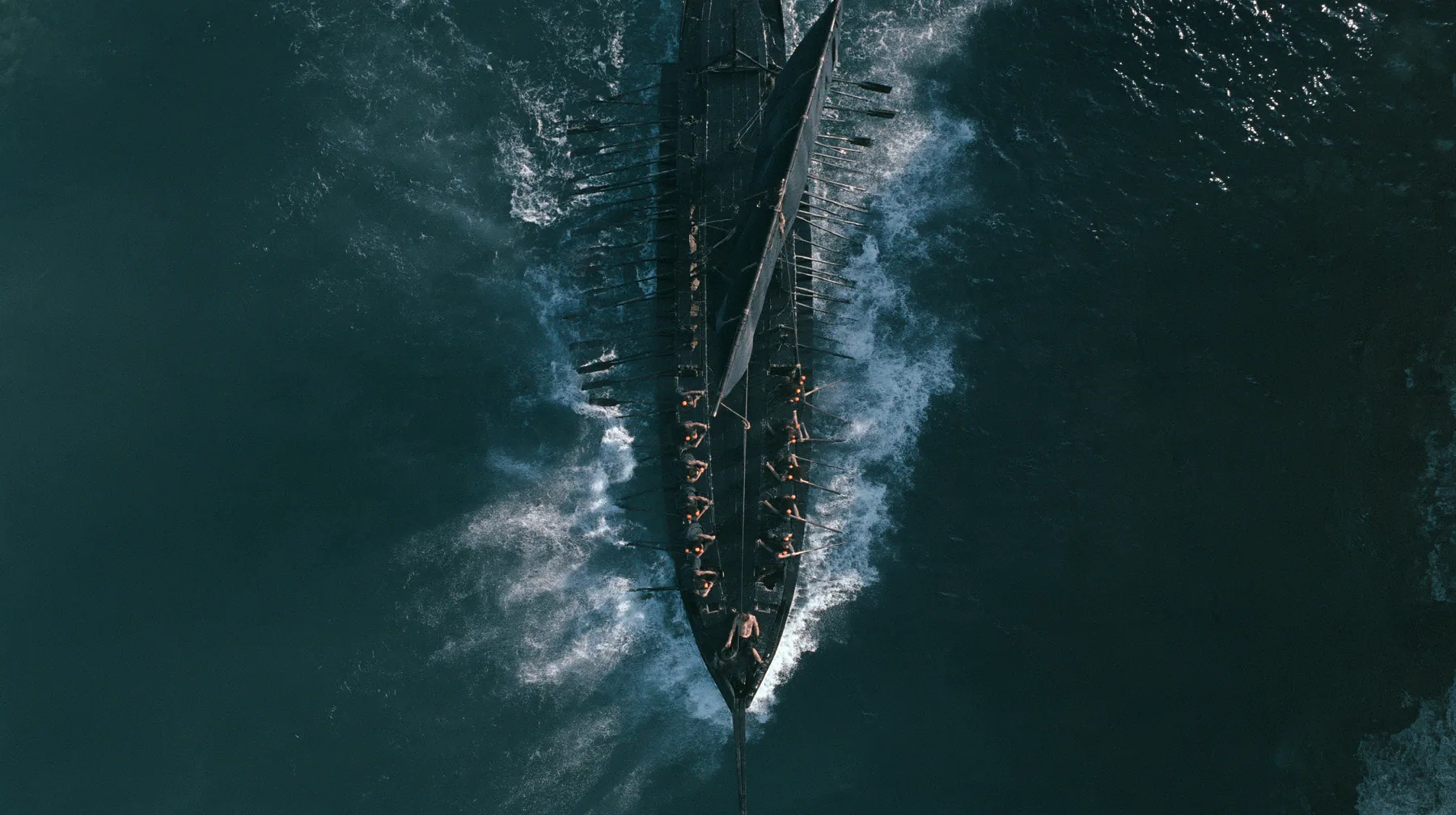 straight-down aerial of a long black ancient Greek galley with a single mast, a square sail and two banks of oars in a narrow strait, half the benches empty, the remaining rowers rowing with orange earplugs, one man…