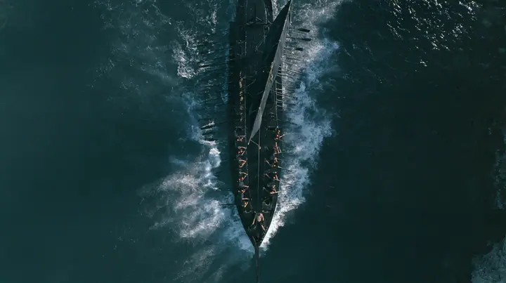 straight-down aerial of a long black ancient Greek galley with a single mast, a square sail and two banks of oars in a narrow strait, half the benches empty, the remaining rowers rowing with orange earplugs, one man…