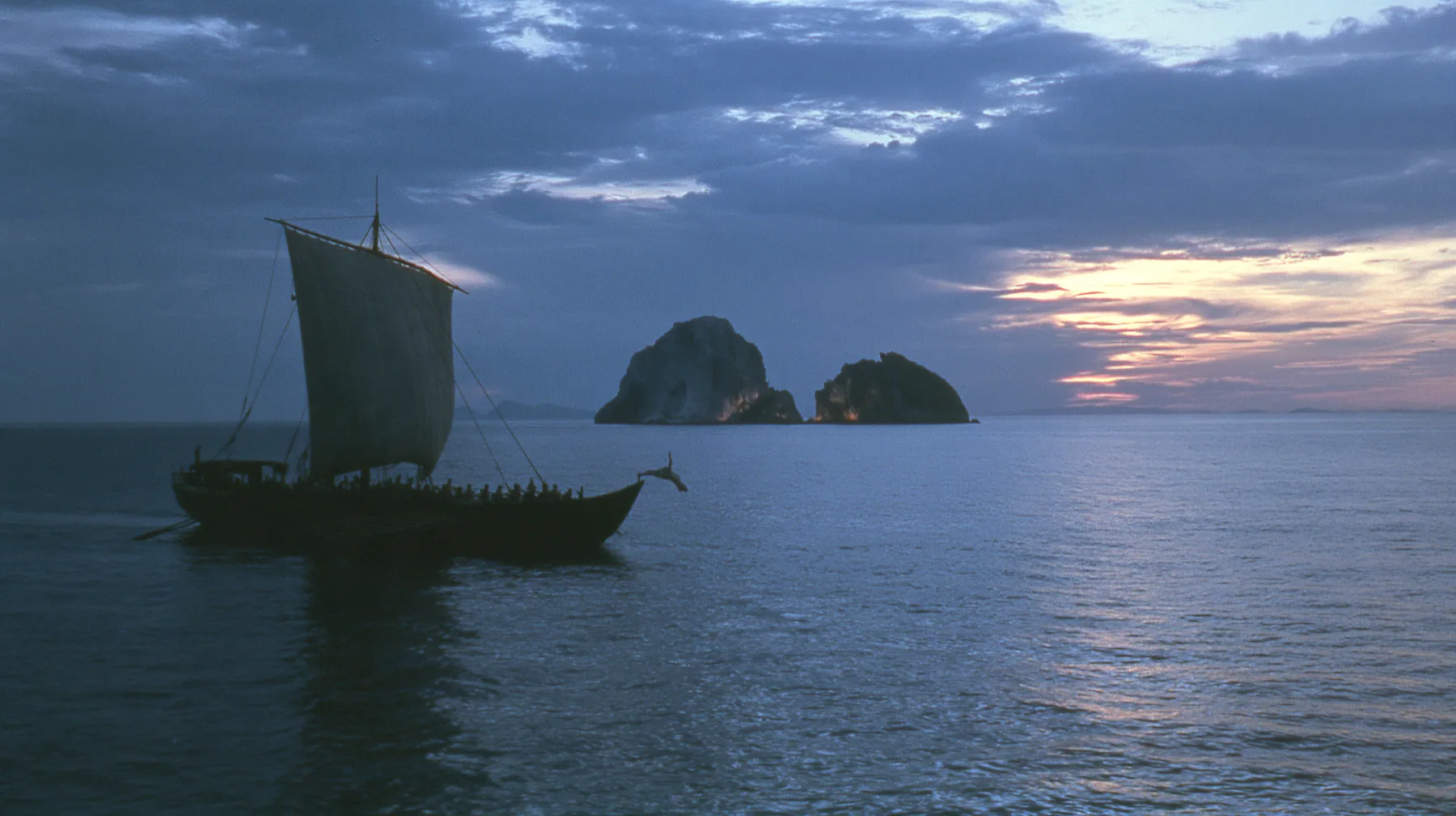a rower in a linen tunic in a joyous swan dive off the rail of a long black ancient Greek galley with a single mast, a square sail and two banks of oars toward distant glowing rocks, deep blue twilight, the last gold…