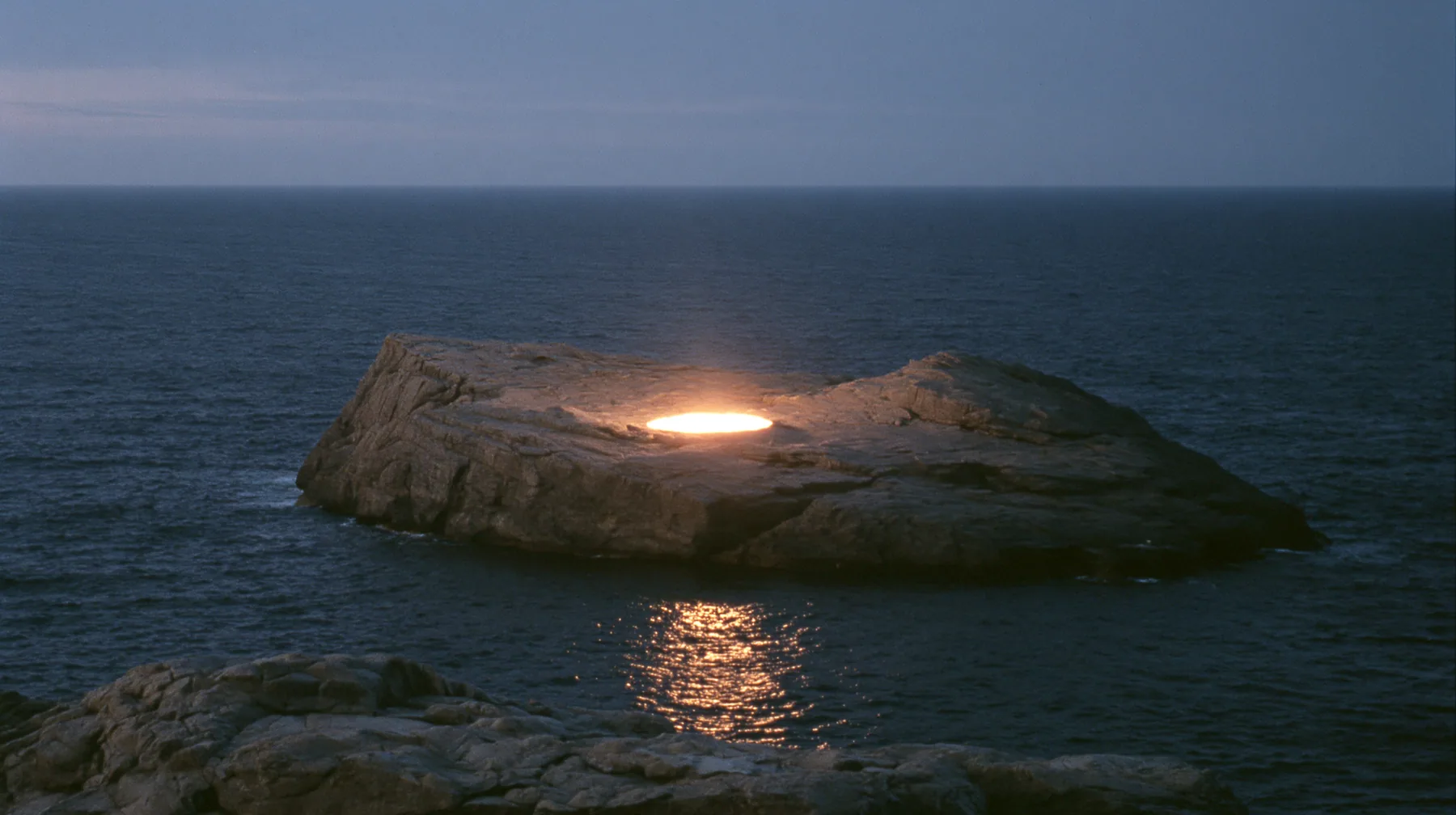 a low black sea rock at twilight with a small circle of warm stage light on it, empty, the dark sea around, deep blue twilight, the last gold on the horizon, glassy sea, cinematic 35mm film still, anamorphic, fine…