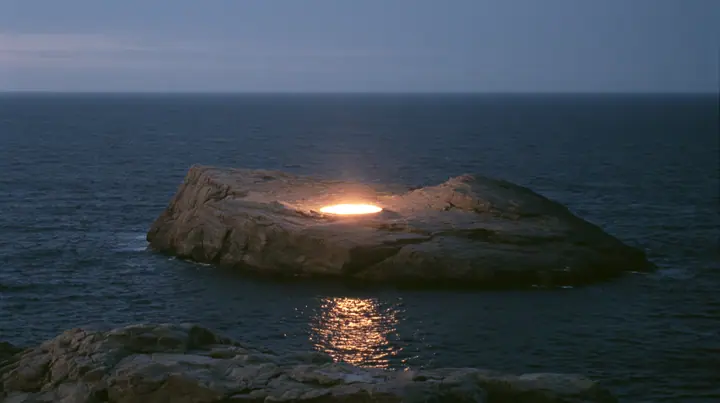 a low black sea rock at twilight with a small circle of warm stage light on it, empty, the dark sea around, deep blue twilight, the last gold on the horizon, glassy sea, cinematic 35mm film still, anamorphic, fine…