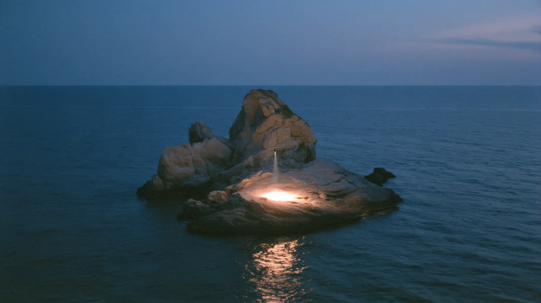 a low black sea rock at twilight with a small circle of warm stage light on it, empty, the dark sea around, deep blue twilight, the last gold on the horizon, glassy sea, cinematic 35mm film still, anamorphic, fine…