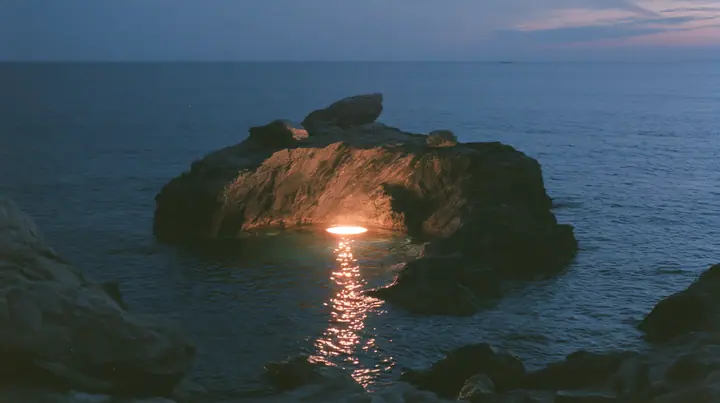 a low black sea rock at twilight with a small circle of warm stage light on it, empty, the dark sea around, deep blue twilight, the last gold on the horizon, glassy sea, cinematic 35mm film still, anamorphic, fine…