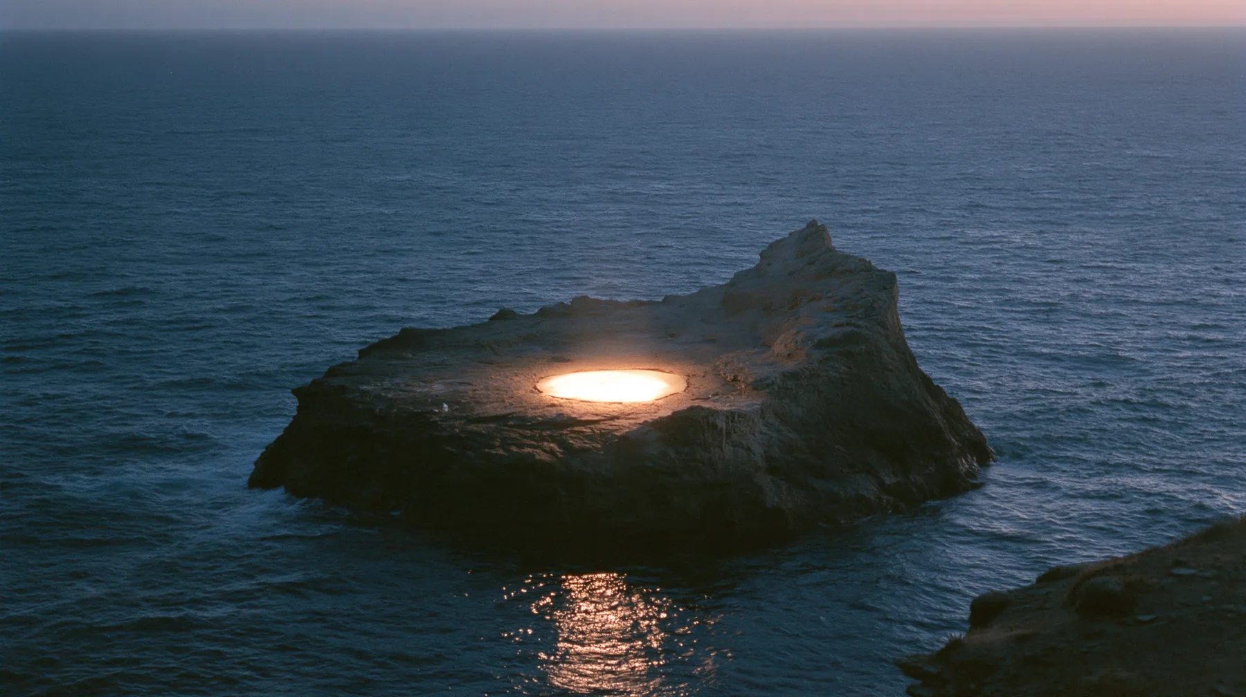 a low black sea rock at twilight with a small circle of warm stage light on it, empty, the dark sea around, deep blue twilight, the last gold on the horizon, glassy sea, cinematic 35mm film still, anamorphic, fine…