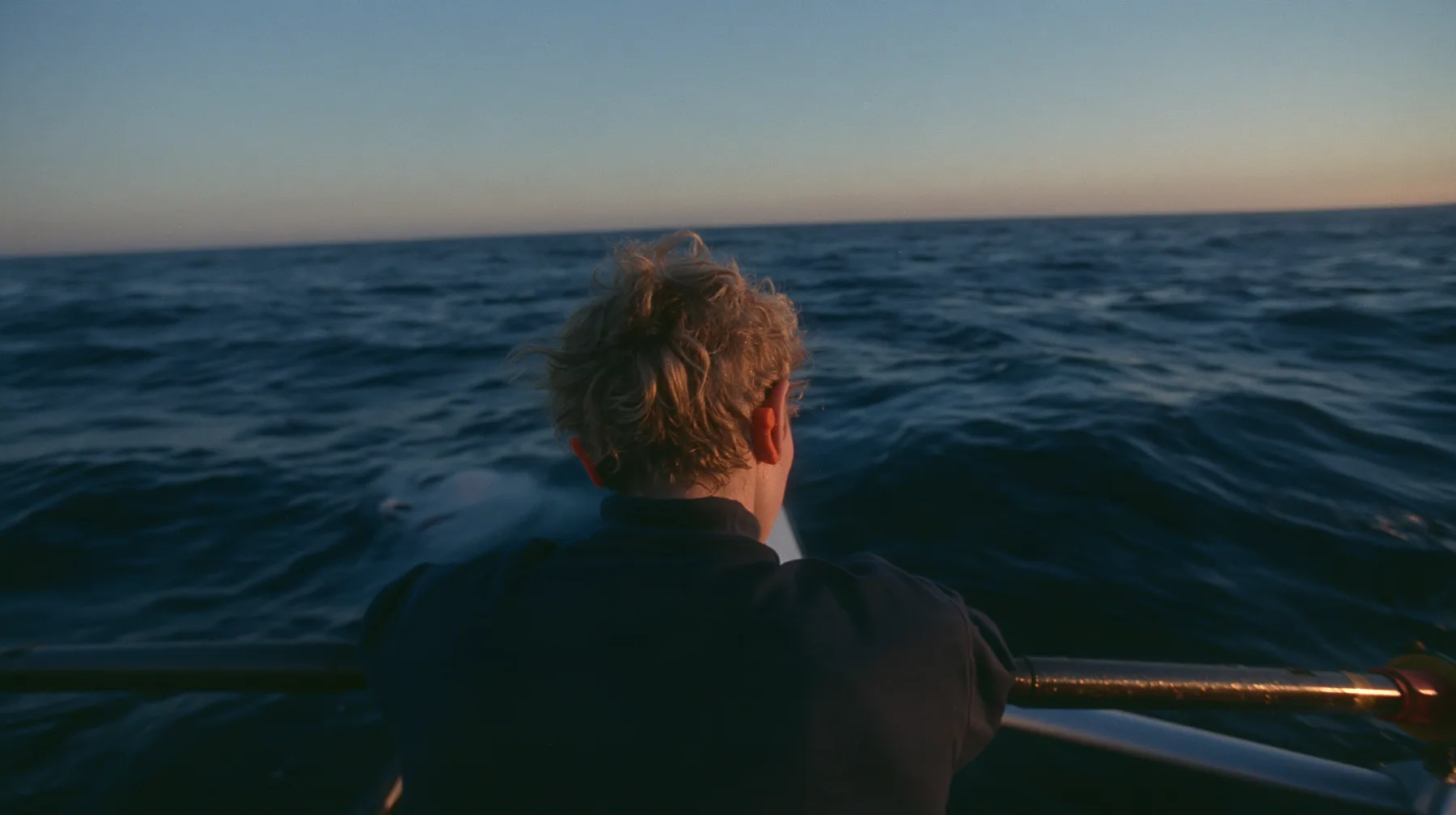over-the-shoulder shot of a rower with orange foam earplugs leaning over the rail and looking down into glassy dark water where a colossal pale woman's face with closed eyes slides past beneath the surface, deep blue…