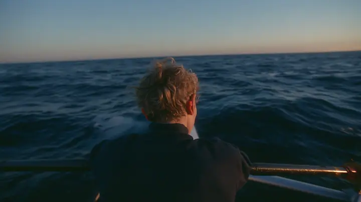 over-the-shoulder shot of a rower with orange foam earplugs leaning over the rail and looking down into glassy dark water where a colossal pale woman's face with closed eyes slides past beneath the surface, deep blue…