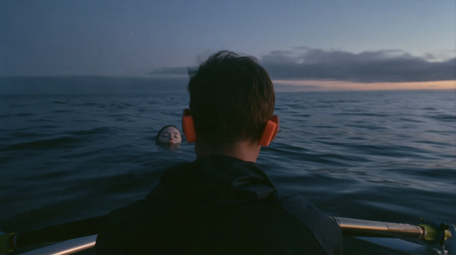 over-the-shoulder shot of a rower with orange foam earplugs leaning over the rail and looking down into glassy dark water where a colossal pale woman's face with closed eyes slides past beneath the surface, deep blue…