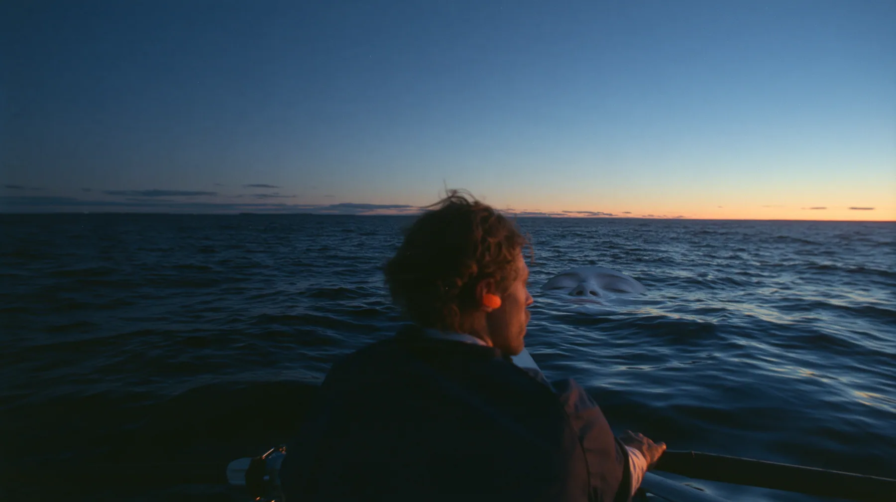 over-the-shoulder shot of a rower with orange foam earplugs leaning over the rail and looking down into glassy dark water where a colossal pale woman's face with closed eyes slides past beneath the surface, deep blue…
