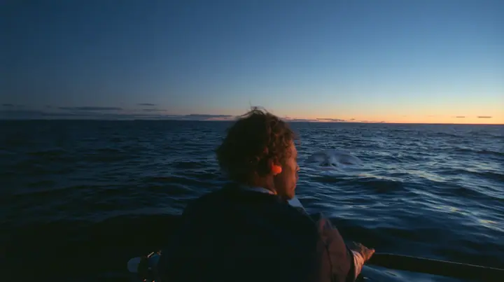 over-the-shoulder shot of a rower with orange foam earplugs leaning over the rail and looking down into glassy dark water where a colossal pale woman's face with closed eyes slides past beneath the surface, deep blue…