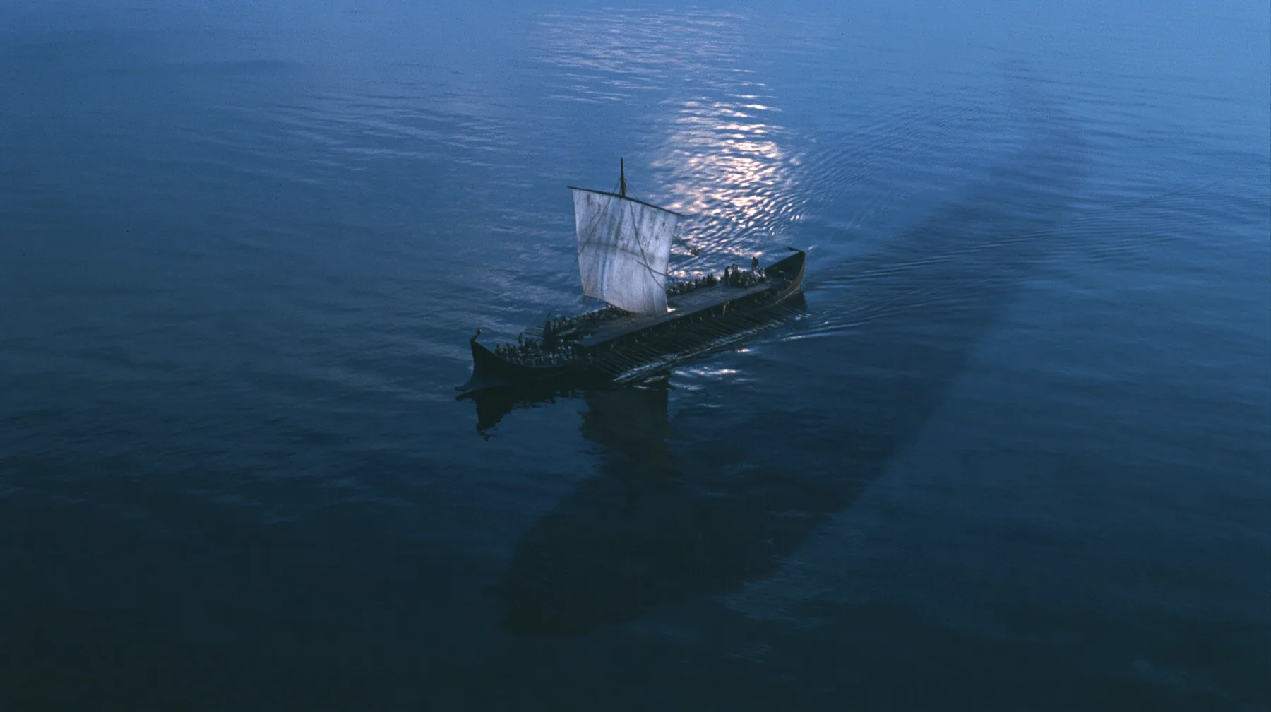 high aerial of a long black ancient Greek galley with a single mast, a square sail and two banks of oars on a darkening sea, beneath it a vast dark shadow ten times the ship's length gliding under the hull, deep blue…