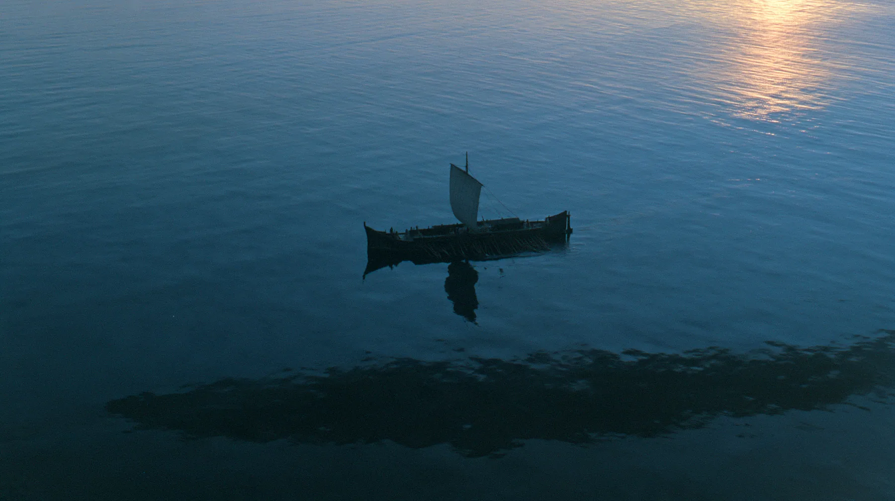 high aerial of a long black ancient Greek galley with a single mast, a square sail and two banks of oars on a darkening sea, beneath it a vast dark shadow ten times the ship's length gliding under the hull, deep blue…