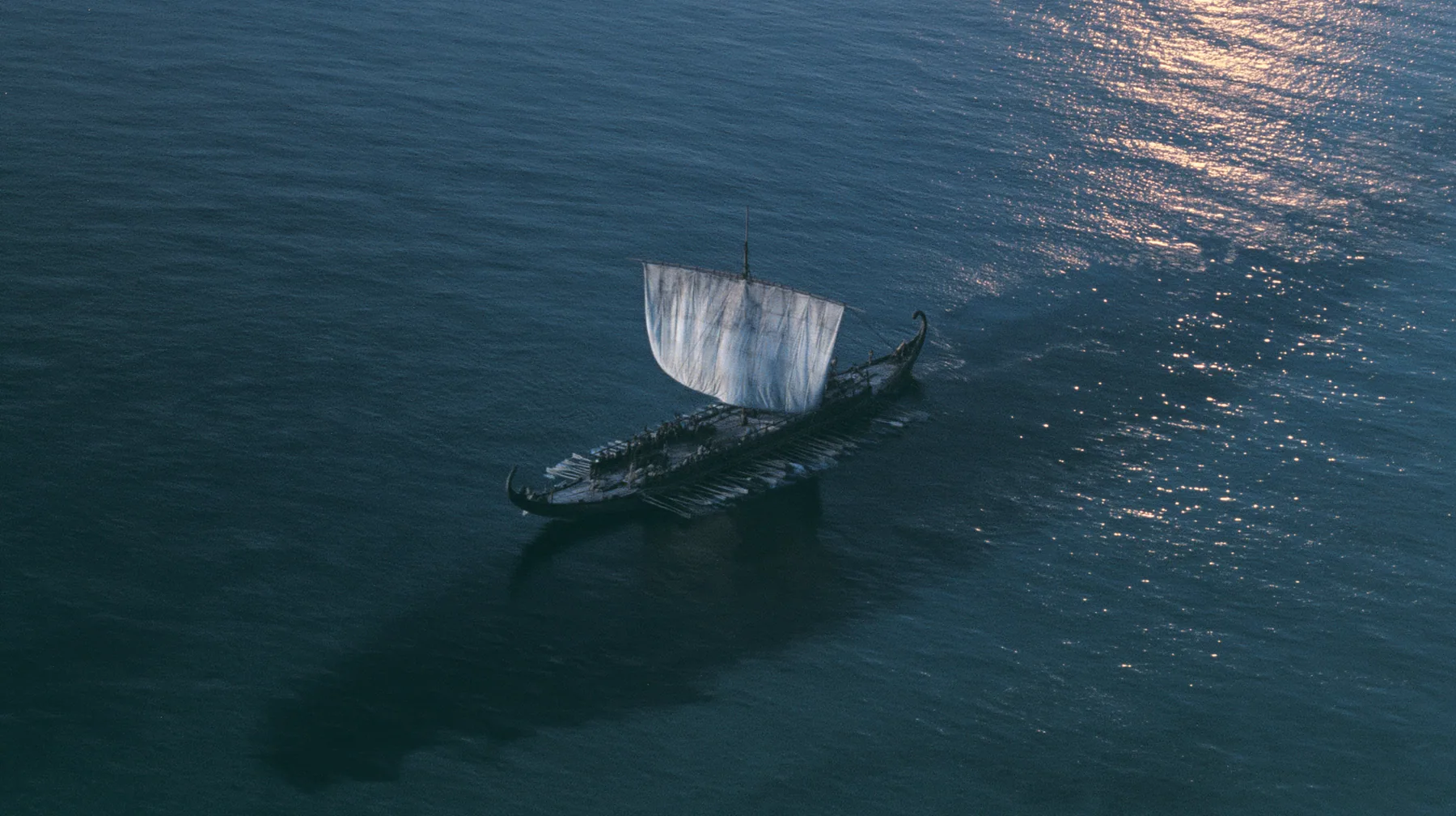high aerial of a long black ancient Greek galley with a single mast, a square sail and two banks of oars on a darkening sea, beneath it a vast dark shadow ten times the ship's length gliding under the hull, deep blue…