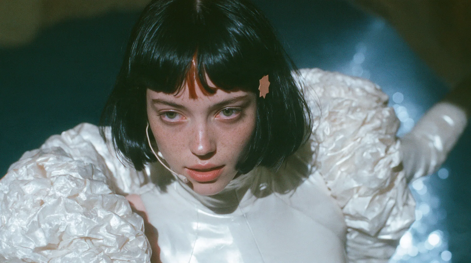 medium shot of a Caucasian American woman in her mid-twenties with pale skin and light freckles, a glossy black blunt jaw-length bob with heavy straight bangs and one clay-orange streak through the bangs, a small…