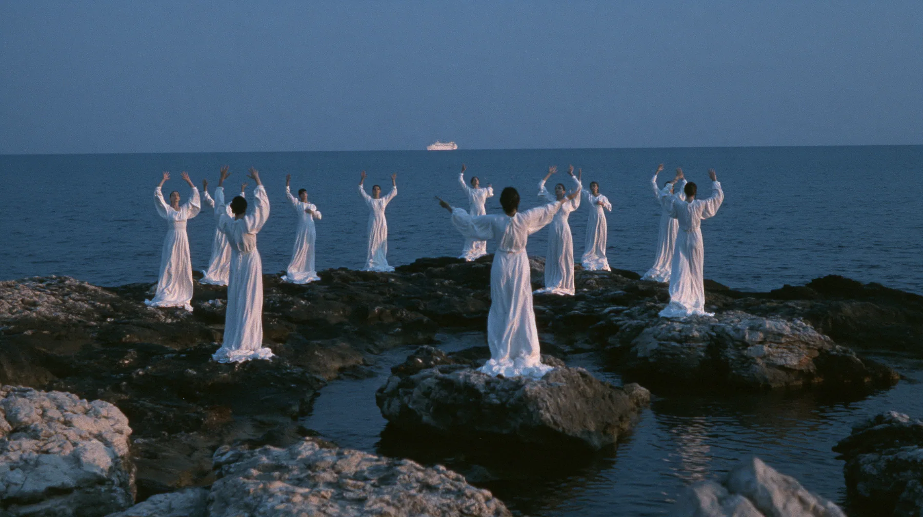 wide shot of hundreds of women in white gowns on black sea rocks raising their arms toward a passing ship, deep blue twilight, the last gold on the horizon, glassy sea, cinematic 35mm film still, anamorphic, fine…