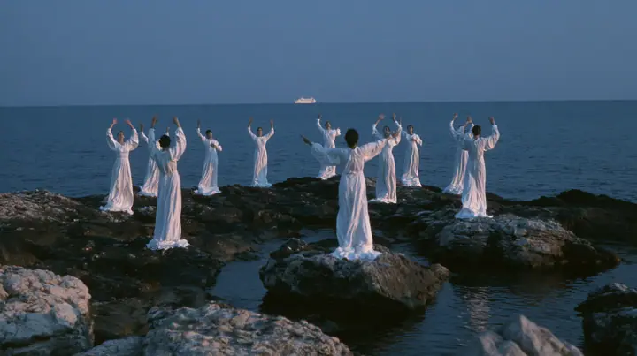 wide shot of hundreds of women in white gowns on black sea rocks raising their arms toward a passing ship, deep blue twilight, the last gold on the horizon, glassy sea, cinematic 35mm film still, anamorphic, fine…