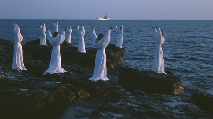 wide shot of hundreds of women in white gowns on black sea rocks raising their arms toward a passing ship, deep blue twilight, the last gold on the horizon, glassy sea, cinematic 35mm film still, anamorphic, fine…