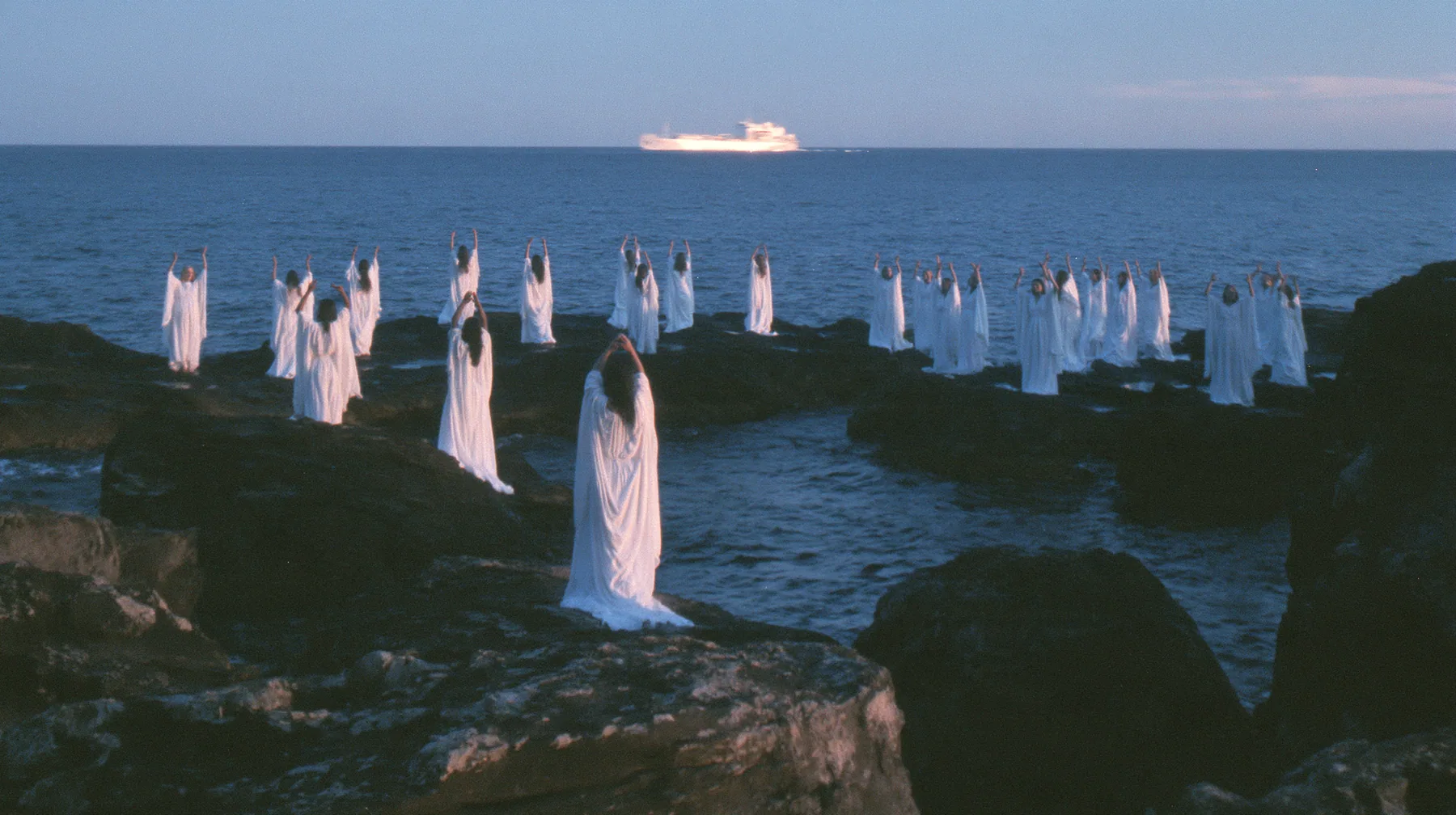 wide shot of hundreds of women in white gowns on black sea rocks raising their arms toward a passing ship, deep blue twilight, the last gold on the horizon, glassy sea, cinematic 35mm film still, anamorphic, fine…