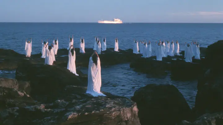 wide shot of hundreds of women in white gowns on black sea rocks raising their arms toward a passing ship, deep blue twilight, the last gold on the horizon, glassy sea, cinematic 35mm film still, anamorphic, fine…
