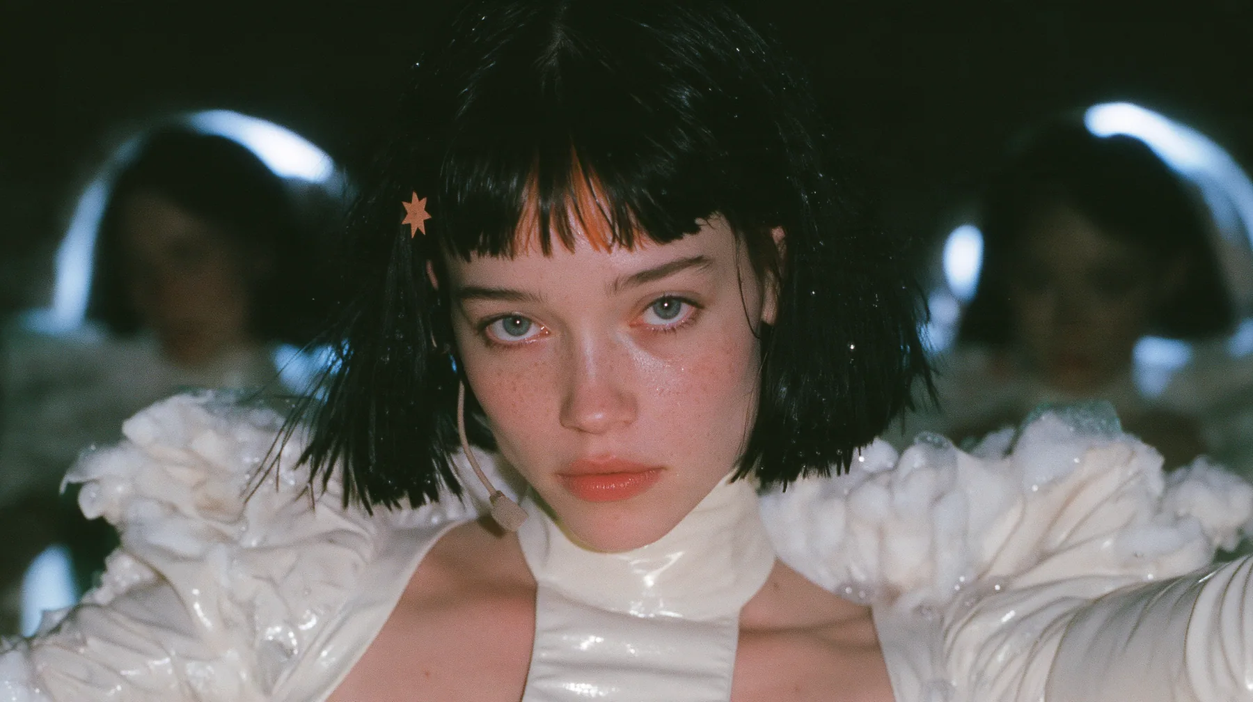 medium shot of a Caucasian American woman in her mid-twenties with pale skin and light freckles, a glossy black blunt jaw-length bob with heavy straight bangs and one clay-orange streak through the bangs, a small…