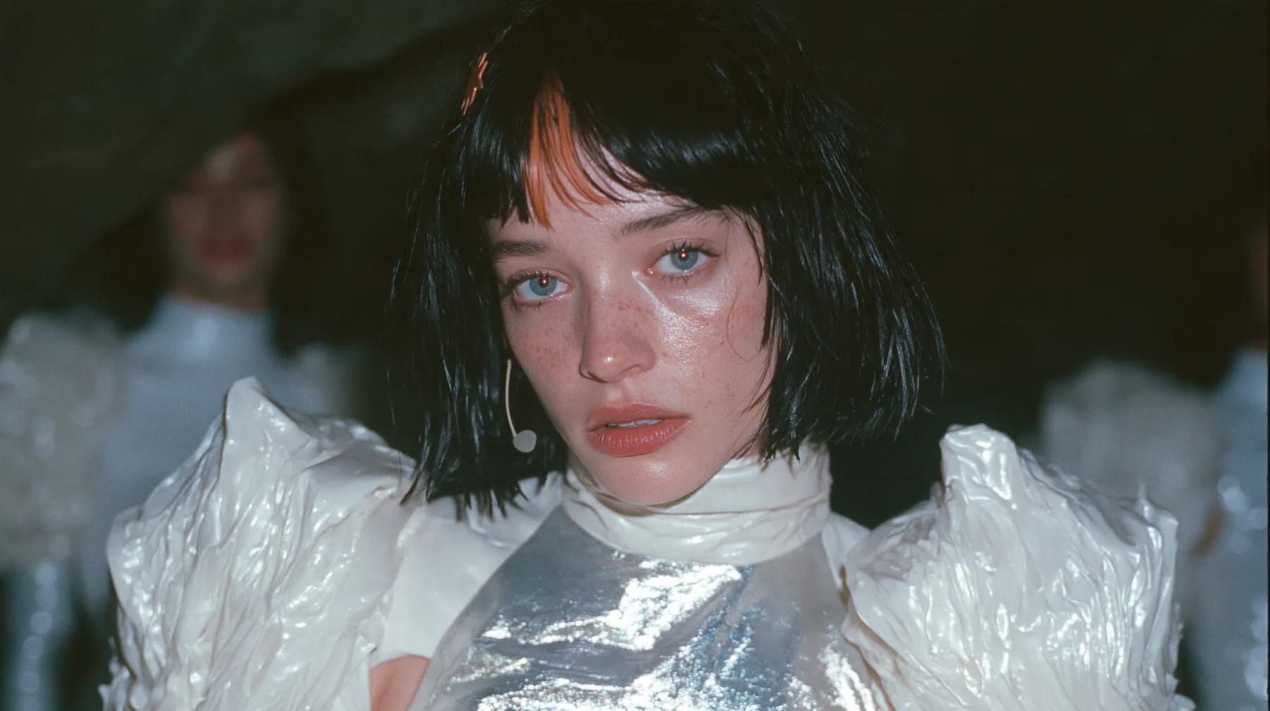 medium shot of a Caucasian American woman in her mid-twenties with pale skin and light freckles, a glossy black blunt jaw-length bob with heavy straight bangs and one clay-orange streak through the bangs, a small…