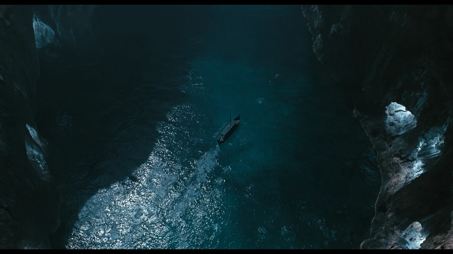 high aerial of the dark water of a narrow sea strait between two cliffs of lit alcoves, tiny swimmers far below drawn toward the cliffs, a long black ancient Greek galley with a single mast, a square sail and two…
