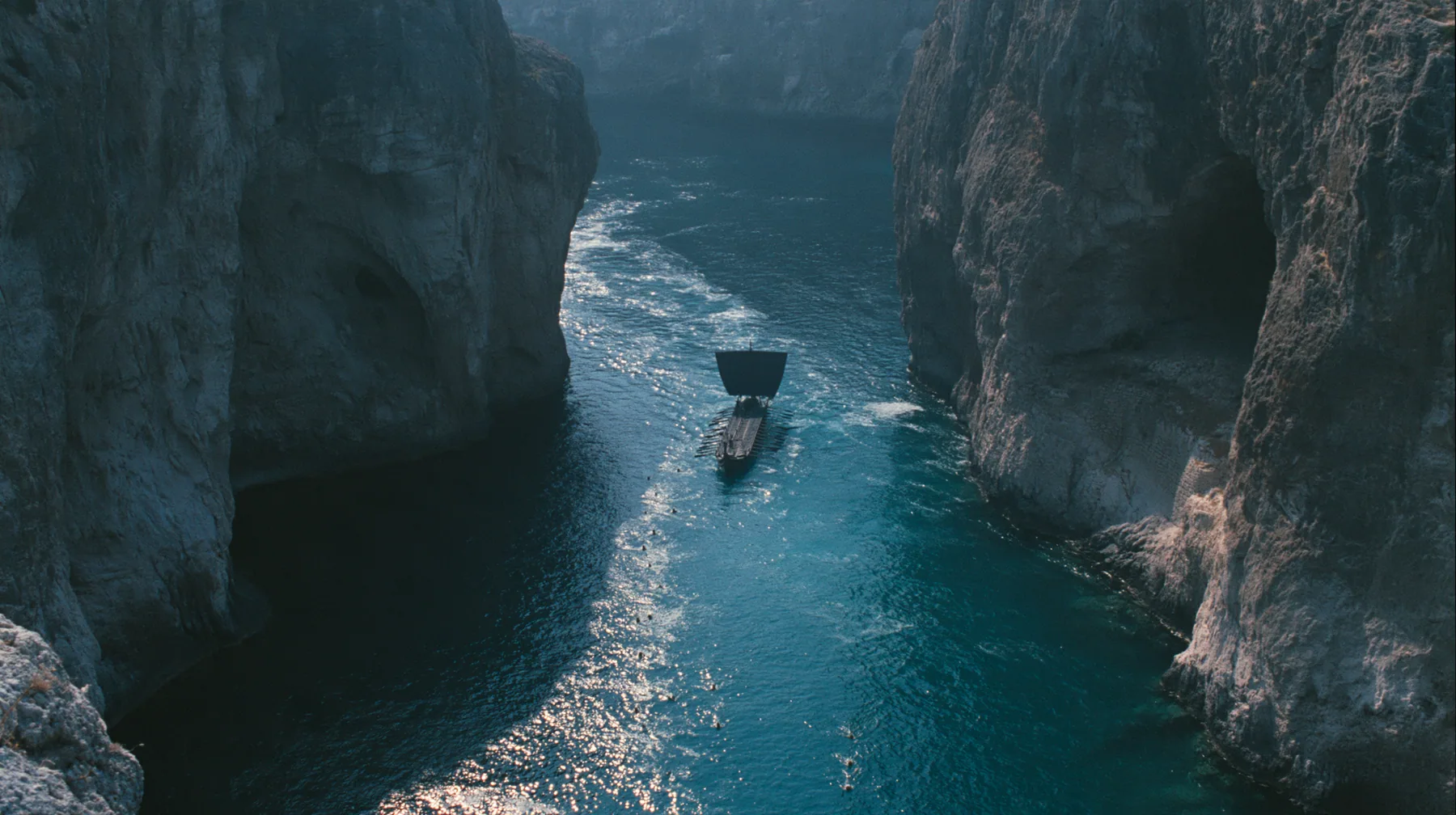 high aerial of the dark water of a narrow sea strait between two cliffs of lit alcoves, tiny swimmers far below drawn toward the cliffs, a long black ancient Greek galley with a single mast, a square sail and two…