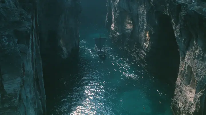 high aerial of the dark water of a narrow sea strait between two cliffs of lit alcoves, tiny swimmers far below drawn toward the cliffs, a long black ancient Greek galley with a single mast, a square sail and two…