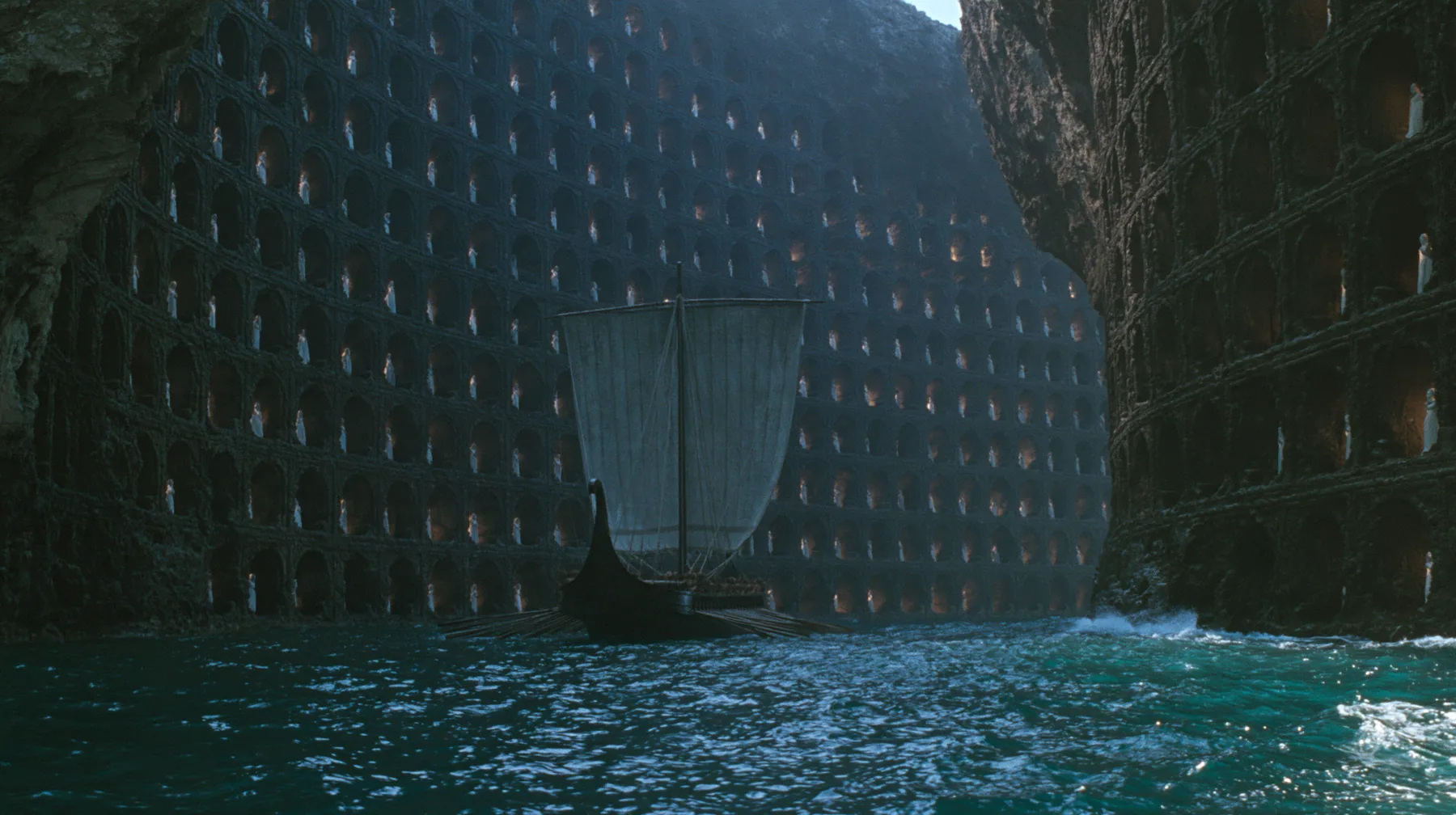 extreme wide shot of a long black ancient Greek galley with a single mast, a square sail and two banks of oars entering a narrow sea strait between two sheer black cliffs hundreds of metres high, the cliff faces…