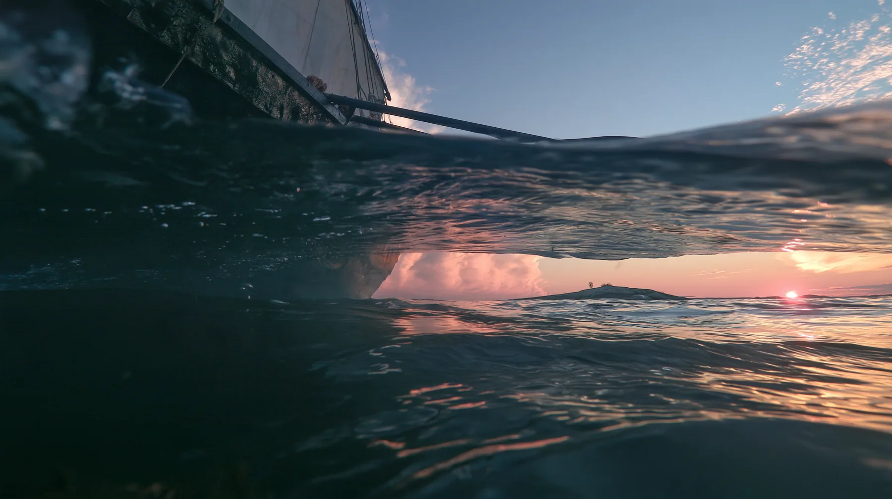 half-underwater split shot at the waterline at twilight, above the black hull of a galley and its dipping oars, below dark water and light shafts, a soft pink-orange glow on a low rock in the distance, no text, no…