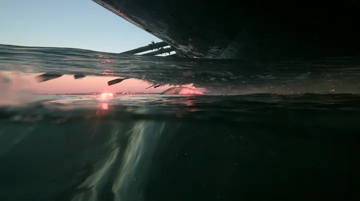 half-underwater split shot at the waterline at twilight, above the black hull of a galley and its dipping oars, below dark water and light shafts, a soft pink-orange glow on a low rock in the distance, no text, no…