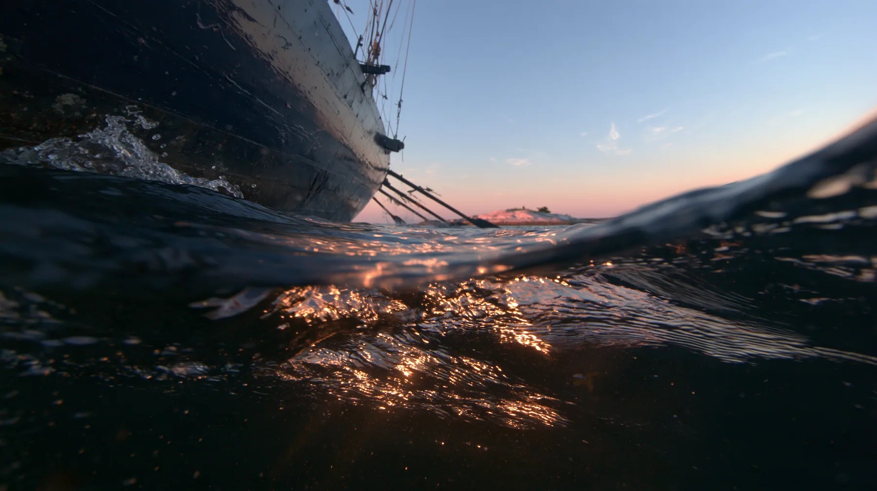 half-underwater split shot at the waterline at twilight, above the black hull of a galley and its dipping oars, below dark water and light shafts, a soft pink-orange glow on a low rock in the distance, no text, no…