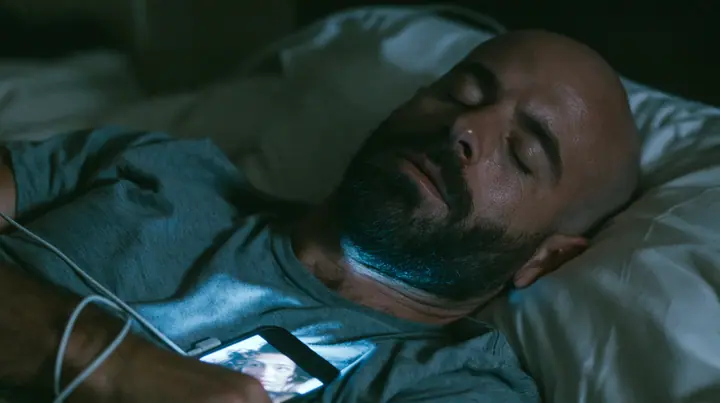 close-up of a broad-shouldered Caucasian man in his early thirties with a shaved head, a thick short dark beard, a square jaw, heavy brows and pale blue eyes, the build of a man who lifts, in a grey t-shirt, asleep…