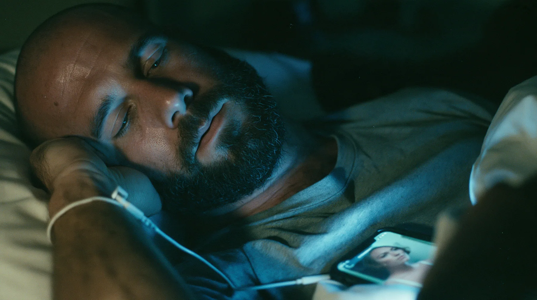 close-up of a broad-shouldered Caucasian man in his early thirties with a shaved head, a thick short dark beard, a square jaw, heavy brows and pale blue eyes, the build of a man who lifts, in a grey t-shirt, asleep…