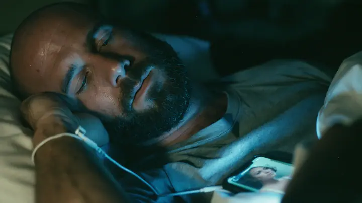 close-up of a broad-shouldered Caucasian man in his early thirties with a shaved head, a thick short dark beard, a square jaw, heavy brows and pale blue eyes, the build of a man who lifts, in a grey t-shirt, asleep…