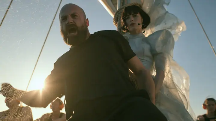 low angle of a broad-shouldered Caucasian man in his early thirties with a shaved head, a thick short dark beard, a square jaw, heavy brows and pale blue eyes, the build of a man who lifts, in a plain black t-shirt…