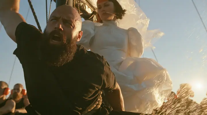 low angle of a broad-shouldered Caucasian man in his early thirties with a shaved head, a thick short dark beard, a square jaw, heavy brows and pale blue eyes, the build of a man who lifts, in a plain black t-shirt…