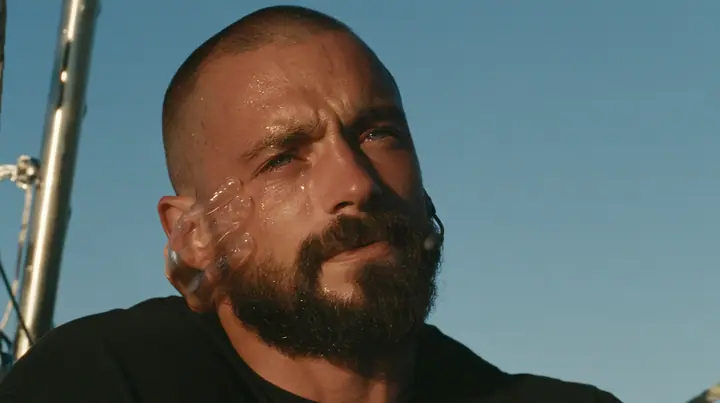 close-up of a broad-shouldered Caucasian man in his early thirties with a shaved head, a thick short dark beard, a square jaw, heavy brows and pale blue eyes, the build of a man who lifts, in a plain black t-shirt…