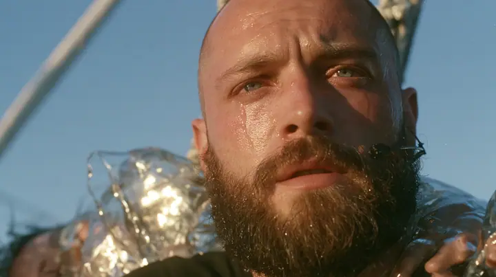 close-up of a broad-shouldered Caucasian man in his early thirties with a shaved head, a thick short dark beard, a square jaw, heavy brows and pale blue eyes, the build of a man who lifts, in a plain black t-shirt…