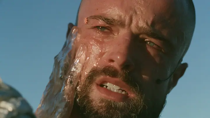 close-up of a broad-shouldered Caucasian man in his early thirties with a shaved head, a thick short dark beard, a square jaw, heavy brows and pale blue eyes, the build of a man who lifts, in a plain black t-shirt…
