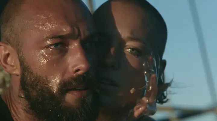 close-up of a broad-shouldered Caucasian man in his early thirties with a shaved head, a thick short dark beard, a square jaw, heavy brows and pale blue eyes, the build of a man who lifts, in a plain black t-shirt…