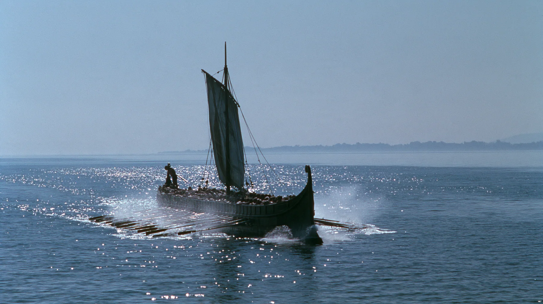 wide low shot of a long black ancient Greek galley with a single mast, a square sail and two banks of oars racing across a sparkling morning sea, oars driving in unison, spray flying, a man tied to the mast, clear…
