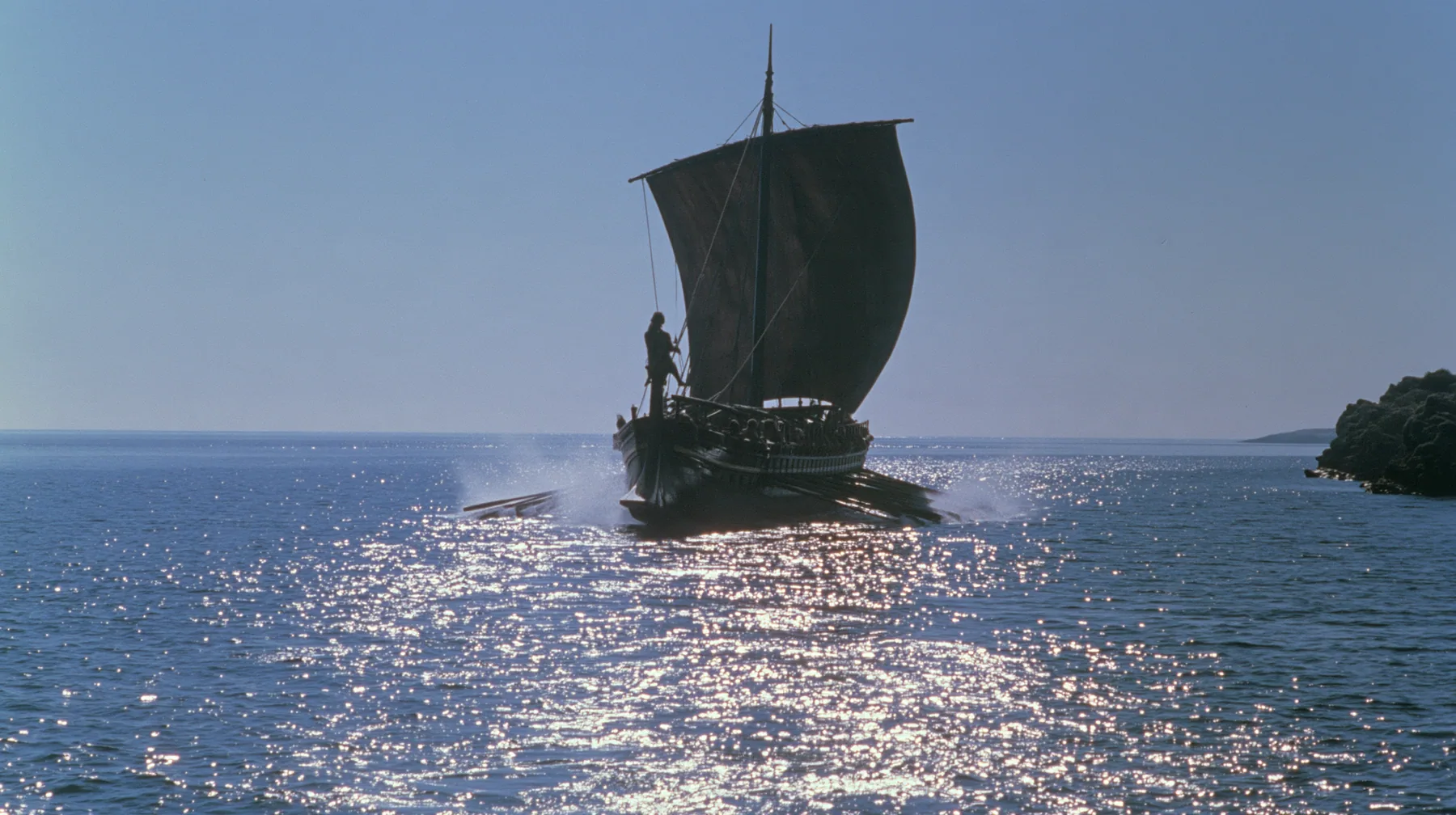 wide low shot of a long black ancient Greek galley with a single mast, a square sail and two banks of oars racing across a sparkling morning sea, oars driving in unison, spray flying, a man tied to the mast, clear…
