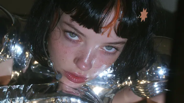 a Caucasian American woman in her mid-twenties with pale skin and light freckles, a glossy black blunt jaw-length bob with heavy straight bangs and one clay-orange streak through the bangs, a small flat clay-orange…