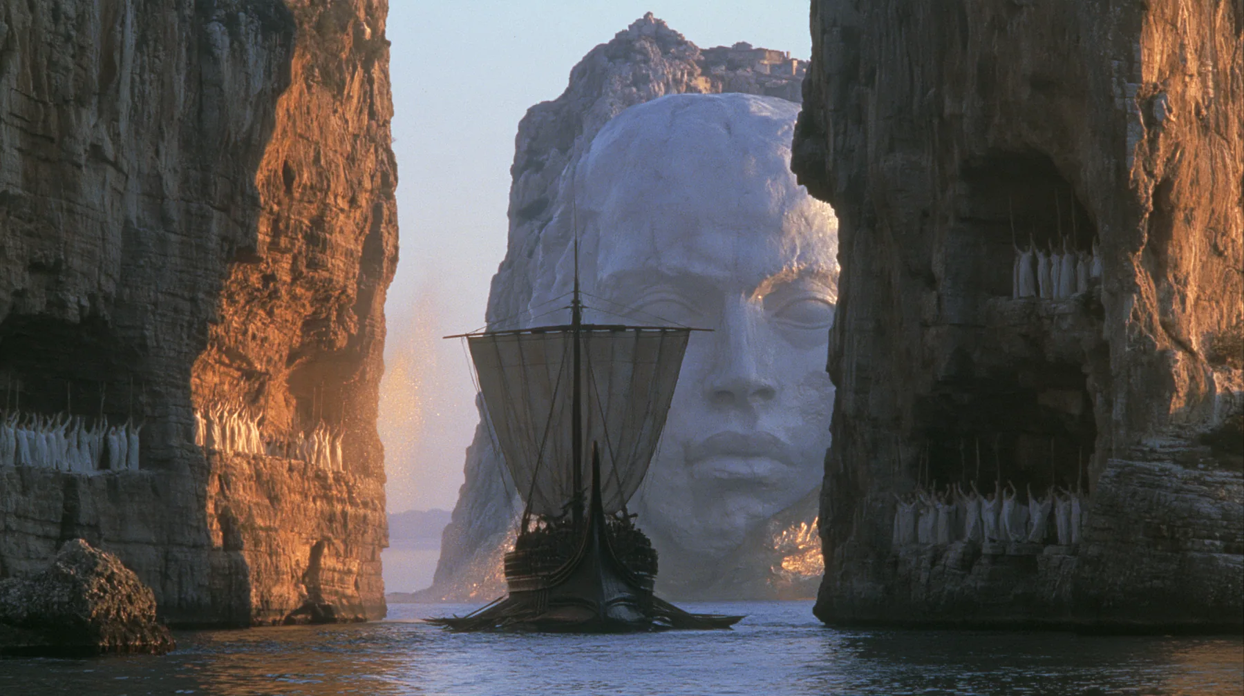 extreme wide shot of a long black ancient Greek galley with a single mast, a square sail and two banks of oars passing between the last lit cliffs of a narrow strait, thousands of women in white raising their arms in…
