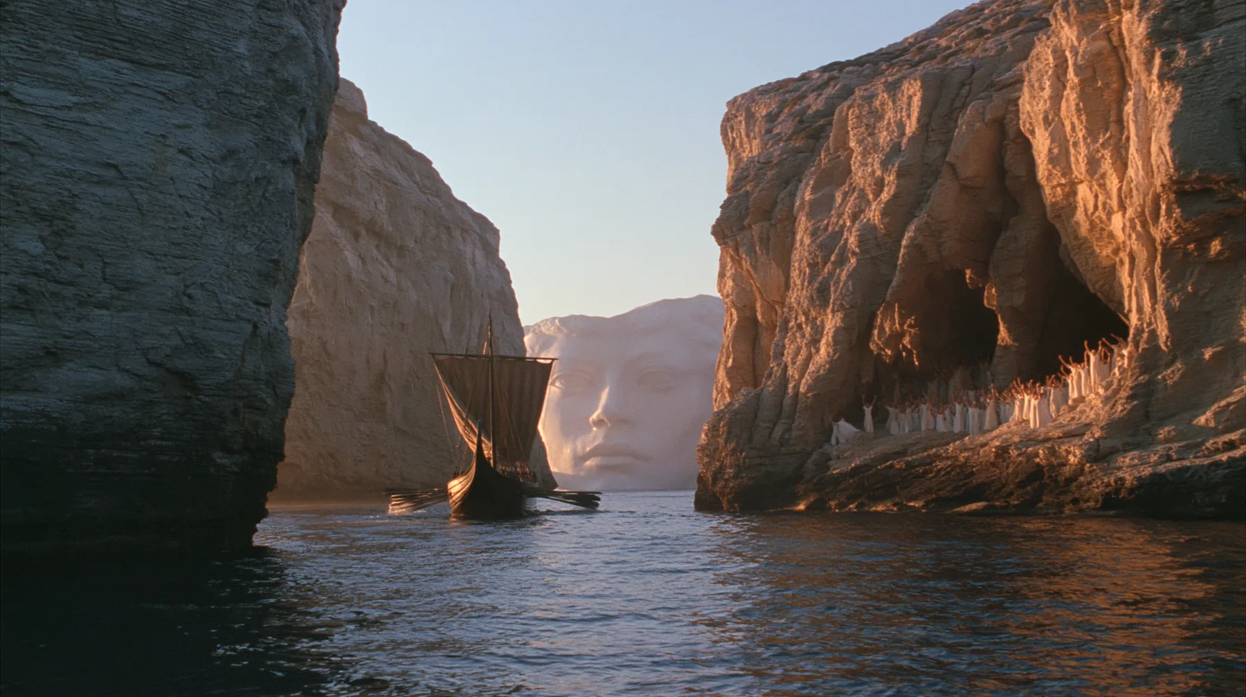extreme wide shot of a long black ancient Greek galley with a single mast, a square sail and two banks of oars passing between the last lit cliffs of a narrow strait, thousands of women in white raising their arms in…