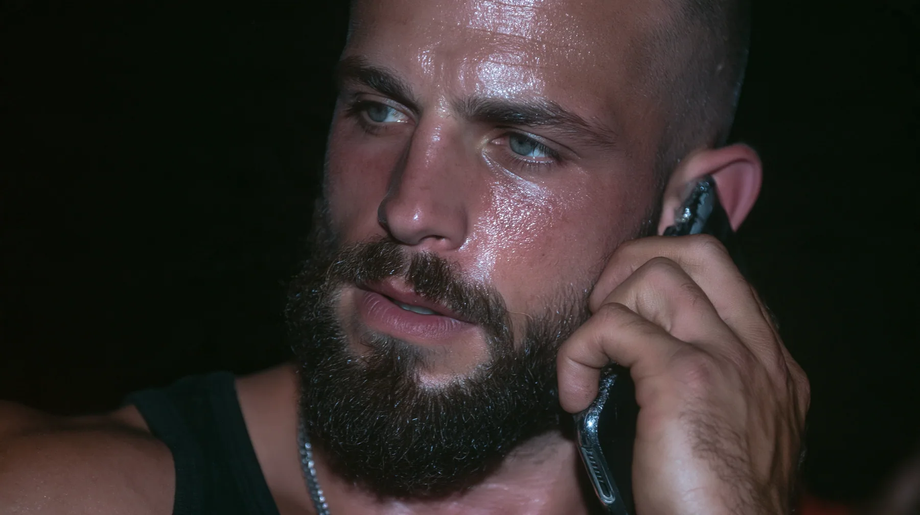 extreme close-up of a phone pressed to the ear of a broad-shouldered Caucasian man in his early thirties with a shaved head, a thick short dark beard, a square jaw, heavy brows and pale blue eyes, the build of a man…