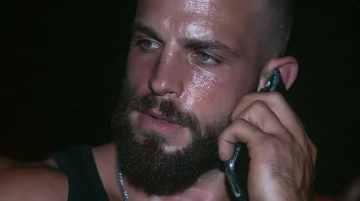 extreme close-up of a phone pressed to the ear of a broad-shouldered Caucasian man in his early thirties with a shaved head, a thick short dark beard, a square jaw, heavy brows and pale blue eyes, the build of a man…